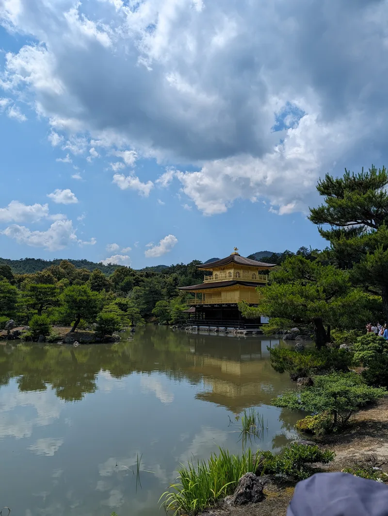Kinkaku-ji Golden Pavilion reflecting in the pond with green trees and blue sky in Kyoto, Japan.