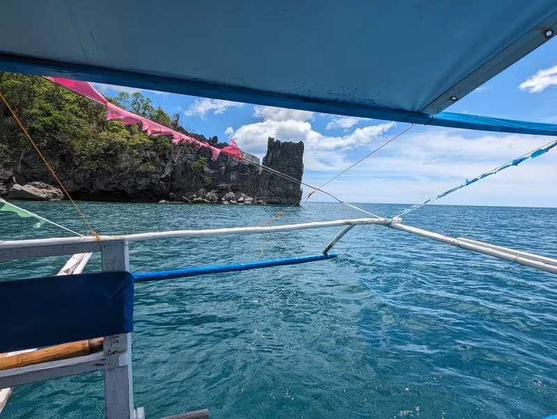 First-person view from the side of a blue bangka boat looking toward a distinctive rocky island with a pink banner draped over the rocks in the clear blue ocean near Sabang, Palawan, Philippines