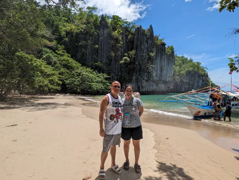 Couple on a sandy beach with limestone cliffs behind them and a traditional Filipino bangka outrigger boat being boarded in the turquoise water to the right at the Subterranean River entrance Sabang, Palawan, Philippines