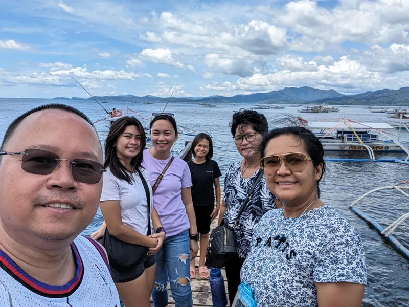 Group selfie on a wooden pier with a man in the foreground and five women behind him smiling with numerous bangka outrigger boats docked in the background at the Subterranean River park entrance Sabang, Palawan, Philippines