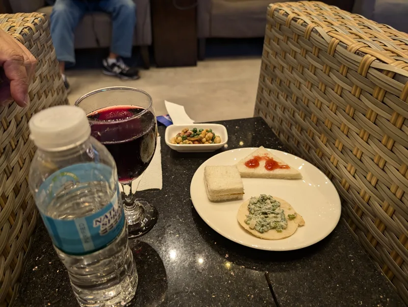 Small plate of lounge snacks with a sandwich, pastry, and nuts alongside a glass of red wine and bottled water at Manila Airport, Philippines.
