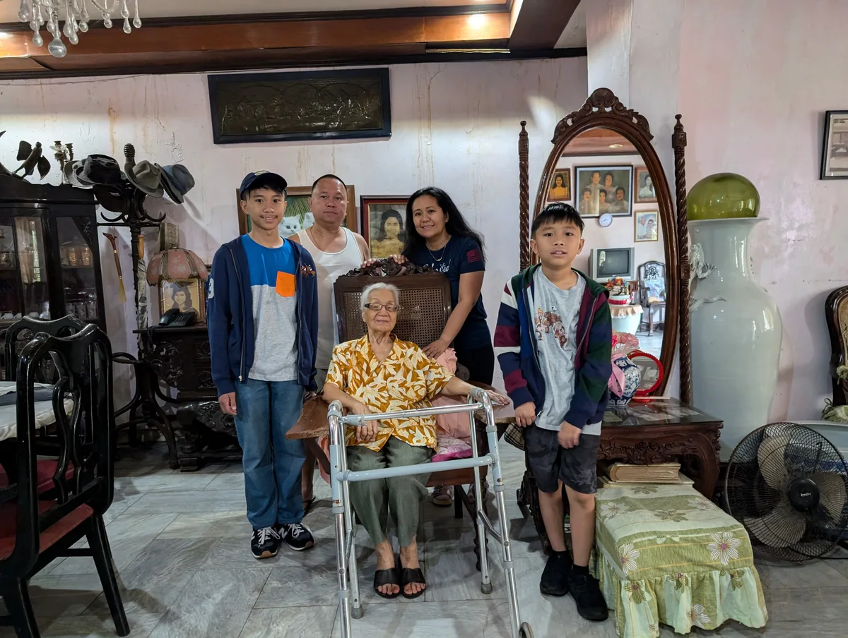 Multi-generational Filipino family group photo in a warmly lit living room in Liliw, Laguna, Philippines, with an elderly woman seated at the center surrounded by family members.