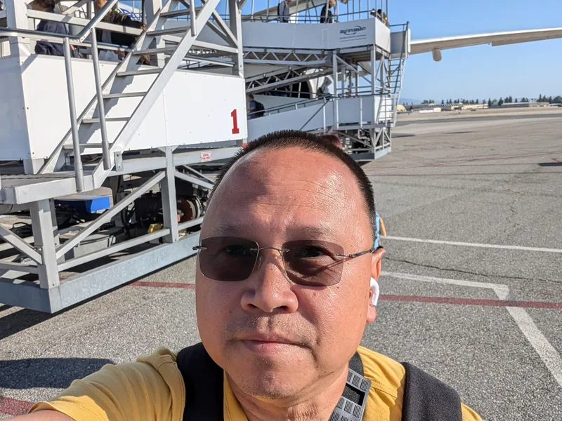 Man in sunglasses taking a selfie on an airport tarmac with boarding stairs and an airplane wing visible behind him, departing Taipei for Ontario, California.