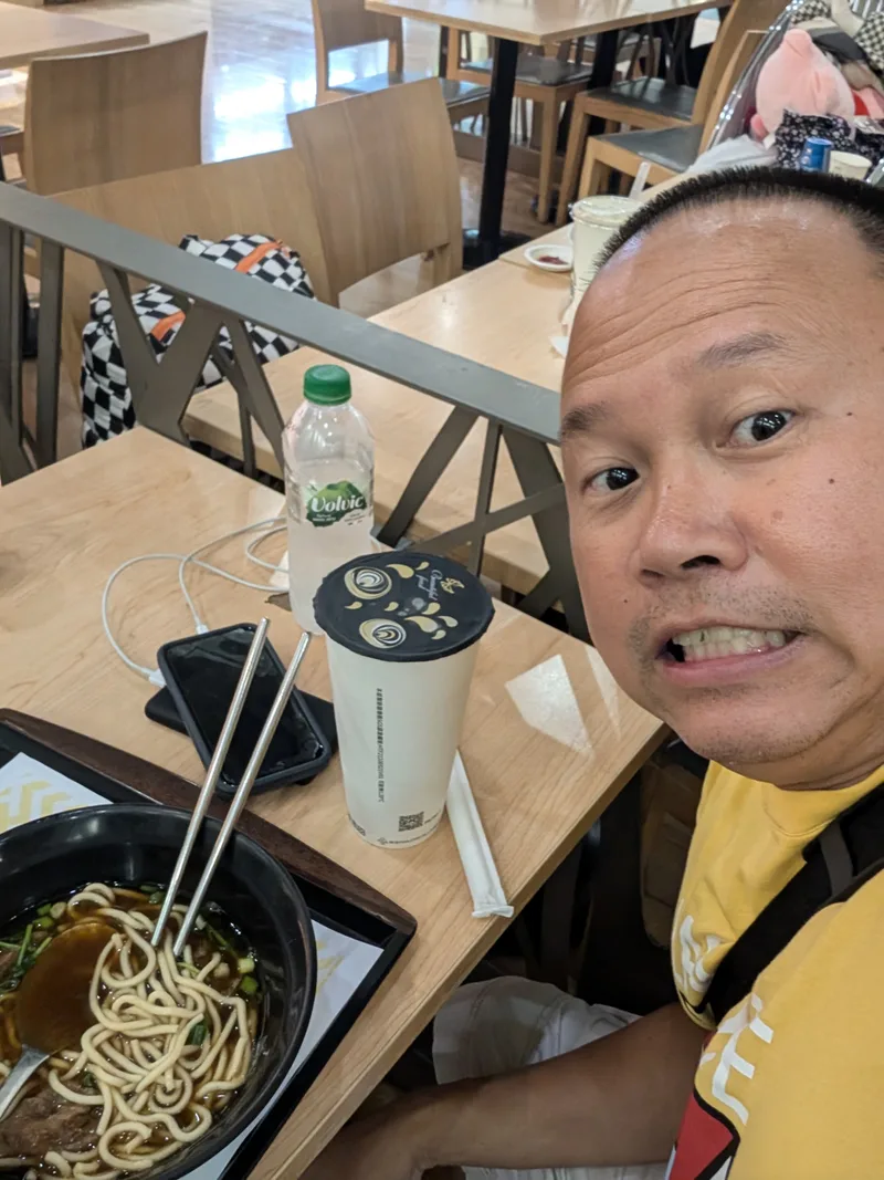 Man taking a selfie at a restaurant table with a bowl of Taiwanese beef noodles and a drink at Taiwan Taoyuan International Airport.
