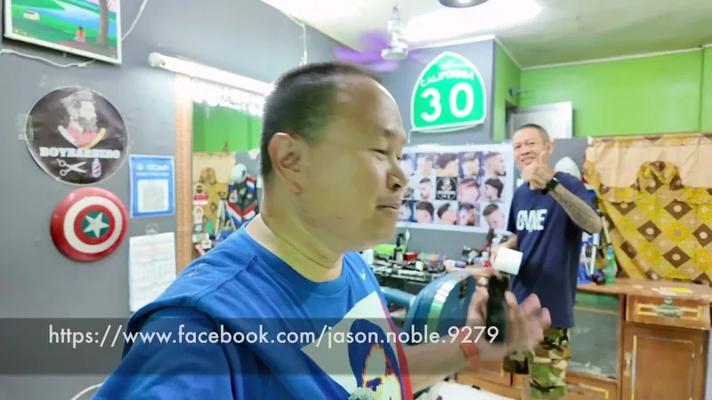 Man in blue shirt in a barber shop, another man in dark shirt and decorations in background.