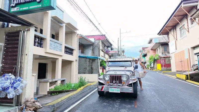 Man in white tank top standing next to a unique silver jeepney on a wet street in the Philippines.