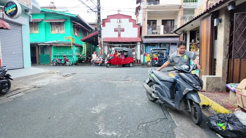 Man pushing a black scooter on a street with a red tricycle and colorful buildings in the Philippines.