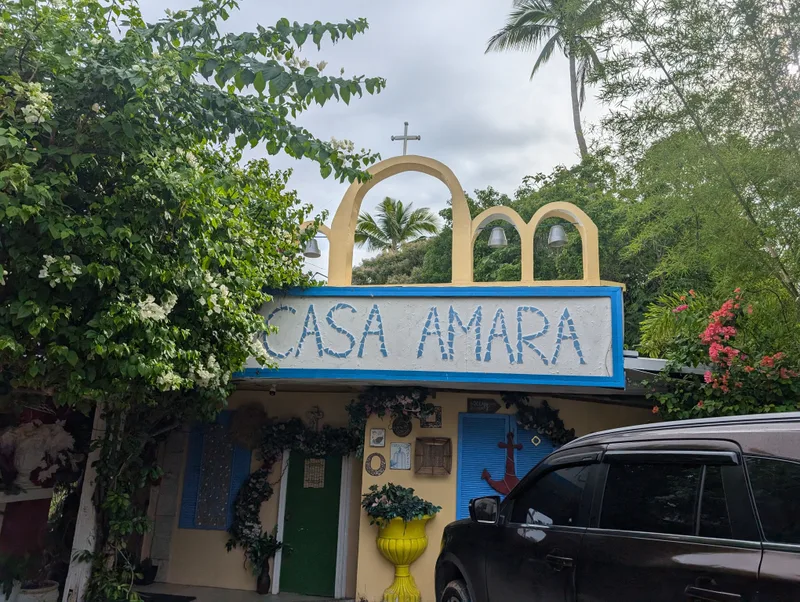 Casa Amara resort entrance with a stone arch gate featuring bells and a cross on top, surrounded by lush green trees and flowering bushes in Batangas, Philippines.