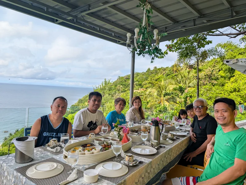 Family group seated at an outdoor dining table with a panoramic ocean and green hill view at Casa Amara, Batangas, Philippines.