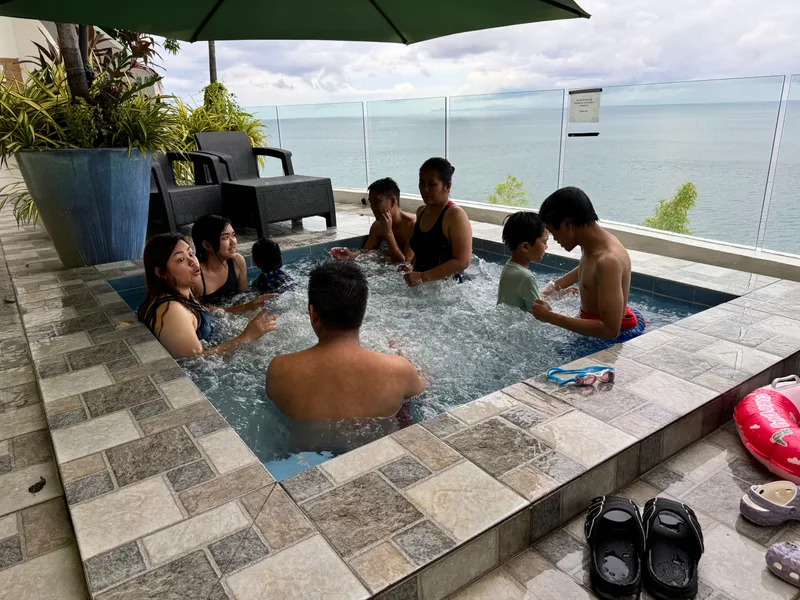Family and friends relaxing in an outdoor jacuzzi with a panoramic ocean view and green umbrella overhead at Casa Amara, Batangas, Philippines.