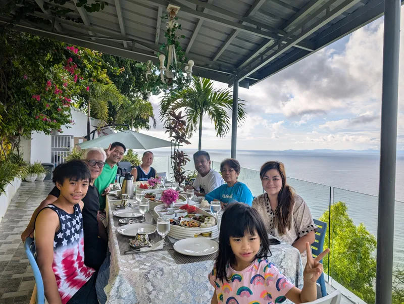 Family dining outdoors at Casa Amara, Batangas, Philippines, with a young girl making a peace sign and the ocean visible behind the group.