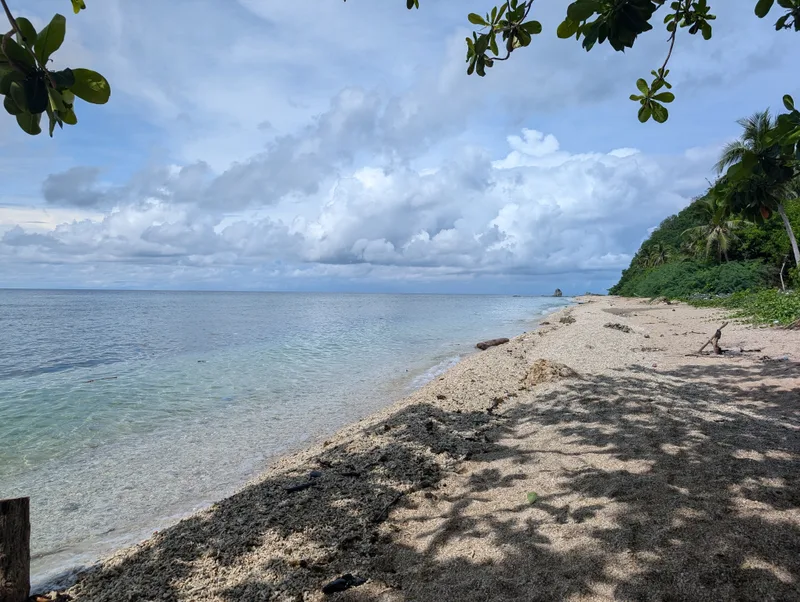 Serene pebble and stone beach with clear turquoise water along the South China Sea coastline, framed by green trees under a partly cloudy sky, in Batangas, Philippines.