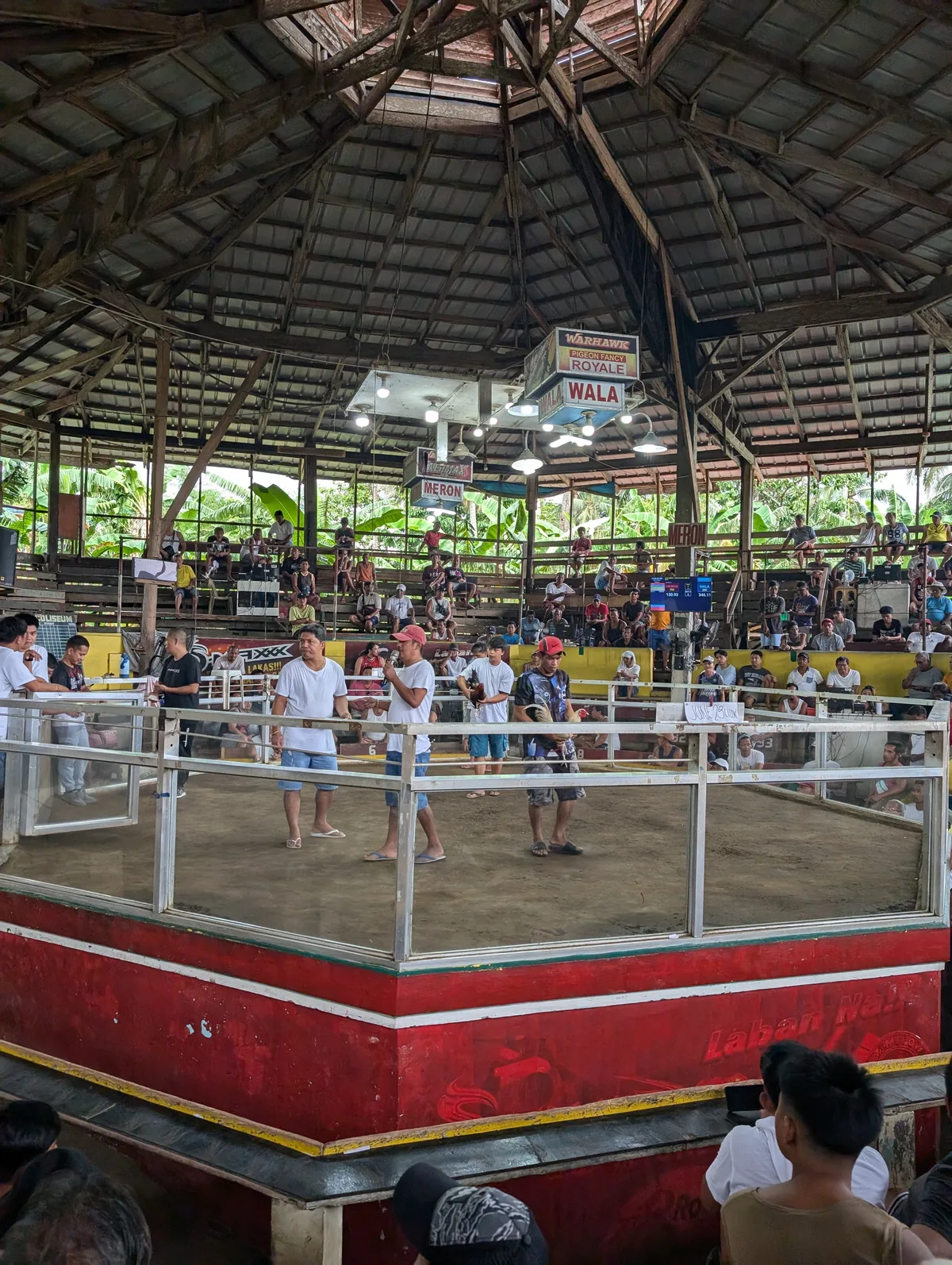 Interior of a circular open-air cockfighting coliseum in Liliw, Laguna, Philippines, with wooden tiered seating and men standing inside the glass-enclosed pit.