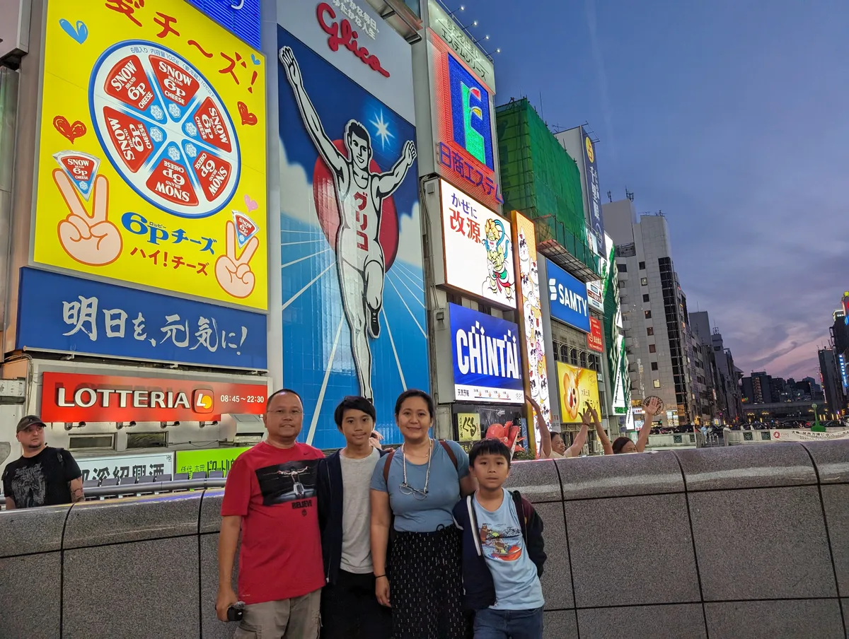 Family of four in front of the Glico Man sign from a wide angle at dusk in Dotonbori, Osaka, Japan.