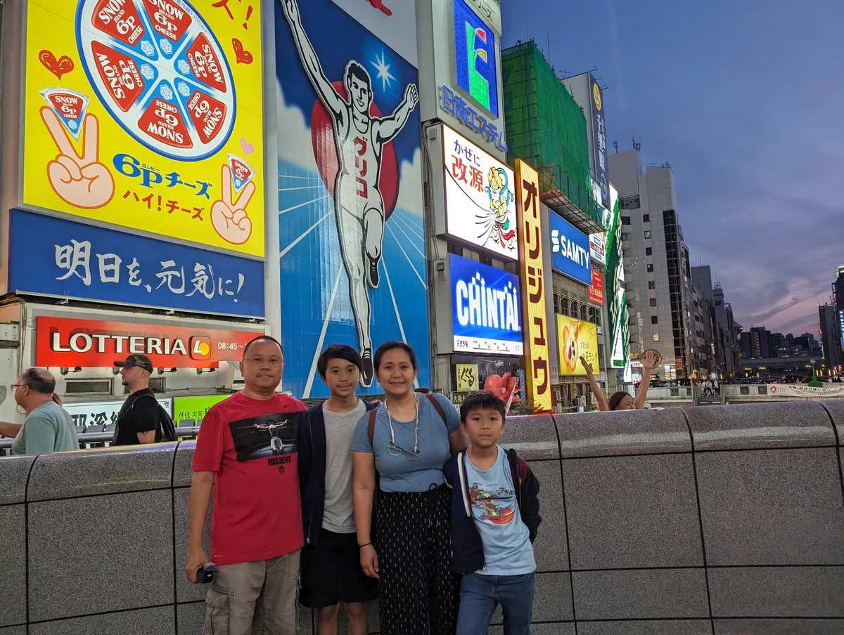 Family of four posing in front of the Glico Man sign with a wide view showing the Dotonbori skyline, Osaka, Japan.