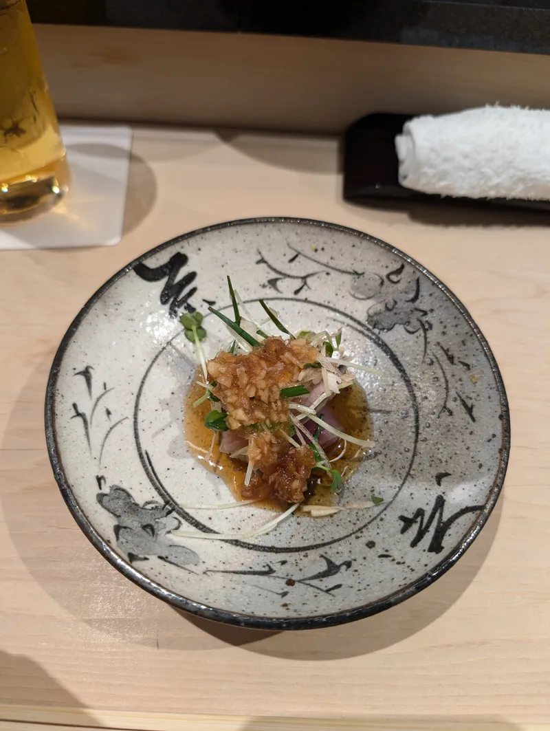 Small dish of raw fish topped with crispy garlic, sprouts, green onions, and dark sauce in a rustic ceramic bowl at an omakase restaurant in Shibuya, Tokyo, Japan.