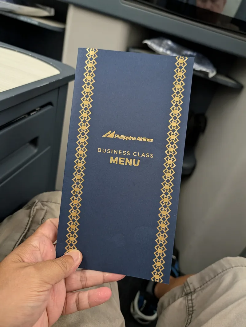 Hand holding a dark blue Philippine Airlines Business Class menu with gold patterned borders in the airplane cabin, Haneda Airport, Tokyo, Japan.