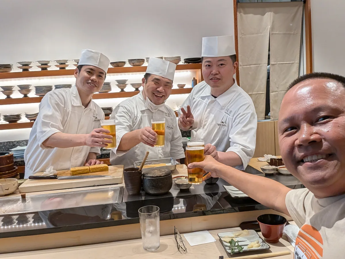 Man toasting Sapporo beers with three smiling Japanese sushi chefs in white uniforms at an omakase counter in Shibuya, Tokyo, Japan.