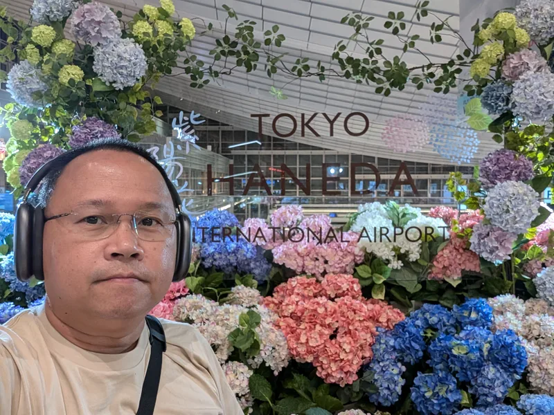 Man wearing headphones and glasses taking a selfie in front of the Tokyo Haneda International Airport sign surrounded by a display of colorful pastel artificial hydrangeas, Tokyo, Japan.