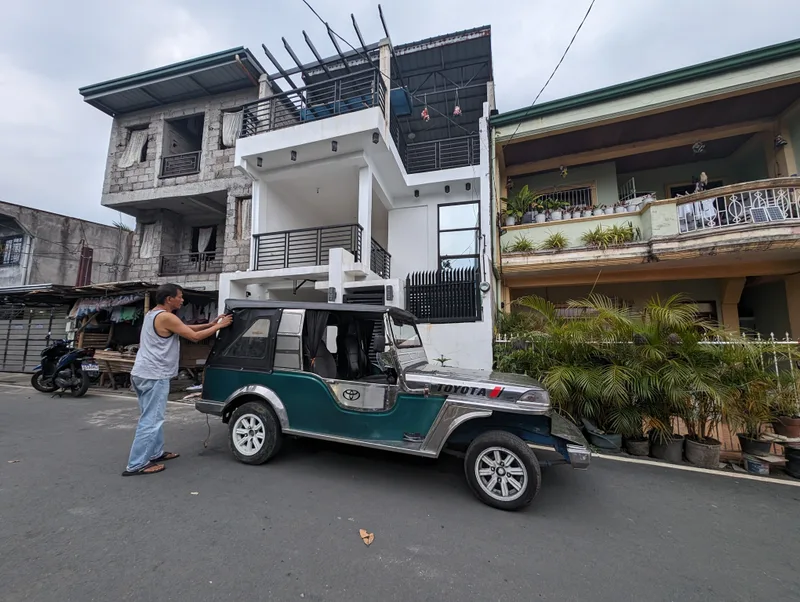 Man in white tank top standing next to his Owner Type Jeep on a street in Liliw, Laguna, Philippines, adjusting the soft top.