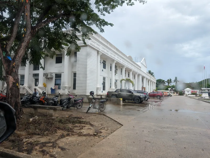 White colonial-style building with columns seen from a car with parked motorcycles and wet ground in Roxas Palawan Philippines