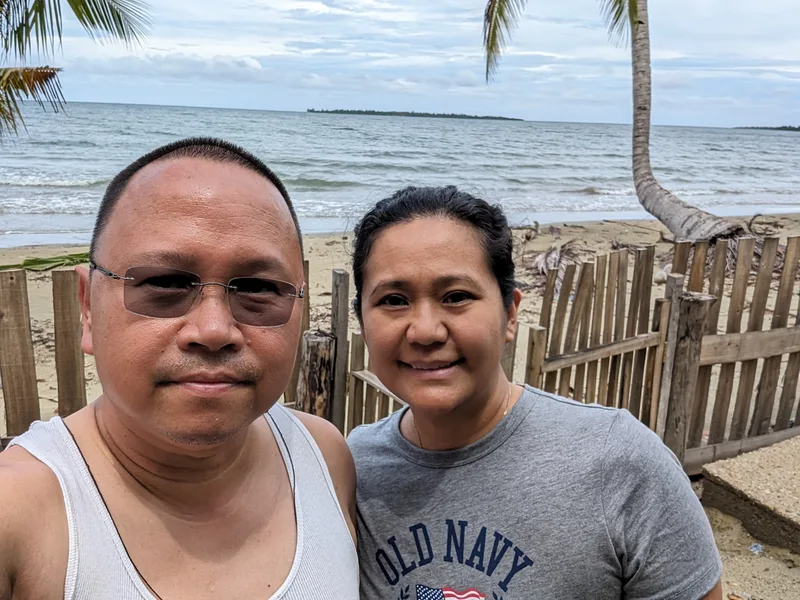 Two people smiling for a selfie on a sandy beach with a wooden fence and the ocean in the background at Henalric resort in Caramay Roxas Palawan