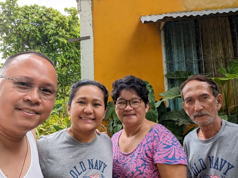 Four people smiling for a group selfie outdoors in front of a bright yellow building with green tropical plants in Roxas Palawan Philippines