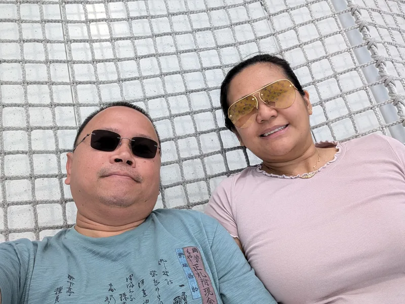 Couple lying on the white mesh net on the Shibuya Sky rooftop in Shibuya, Tokyo, Japan, smiling at the camera, man in dark sunglasses, woman in yellow aviator sunglasses.