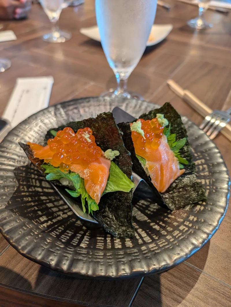 Two salmon and ikura hand rolls wrapped in nori seaweed on a dark textured plate, garnished with green leaves and wasabi, at a restaurant in Shibuya, Tokyo, Japan.