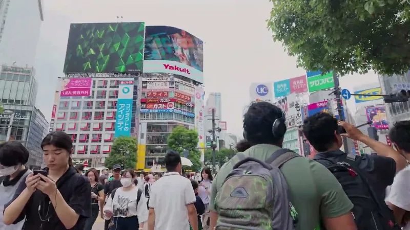 Pedestrians crossing at the famous Shibuya Crossing in Shibuya, Tokyo, Japan, with large digital billboards and tall buildings visible under an overcast sky.