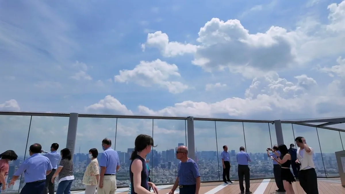 People on the outdoor rooftop observation deck of Shibuya Sky in Tokyo, Japan, with panoramic city views through clean glass barriers under a bright, cloudy sky.