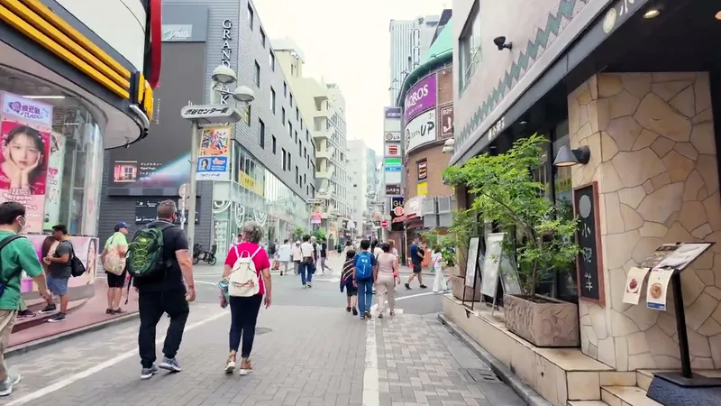 People walking along a narrow street in Shibuya, Tokyo, Japan, lined with shops and restaurants, a large tree on the right.