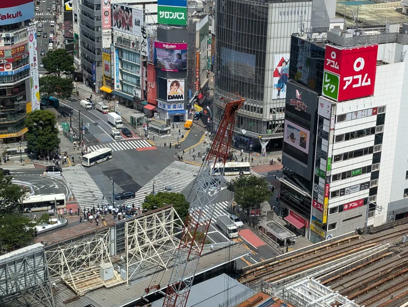 Aerial view of the Shibuya intersection in Tokyo, Japan, from Shibuya Sky, showing pedestrian crosswalks, cars, buses, and surrounding buildings with advertisements.