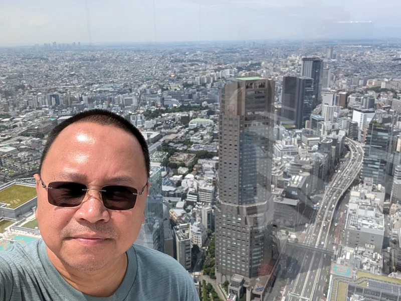 Man in sunglasses taking a selfie at Shibuya Sky with sprawling Tokyo cityscape and tall buildings stretching into the hazy distance behind him, Shibuya, Tokyo, Japan.