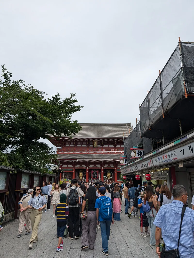 Tourists walking towards Senso-ji Temple along Nakamise-dori market street in Asakusa, Tokyo, Japan, with the temple's main hall visible in the distance under a cloudy sky.