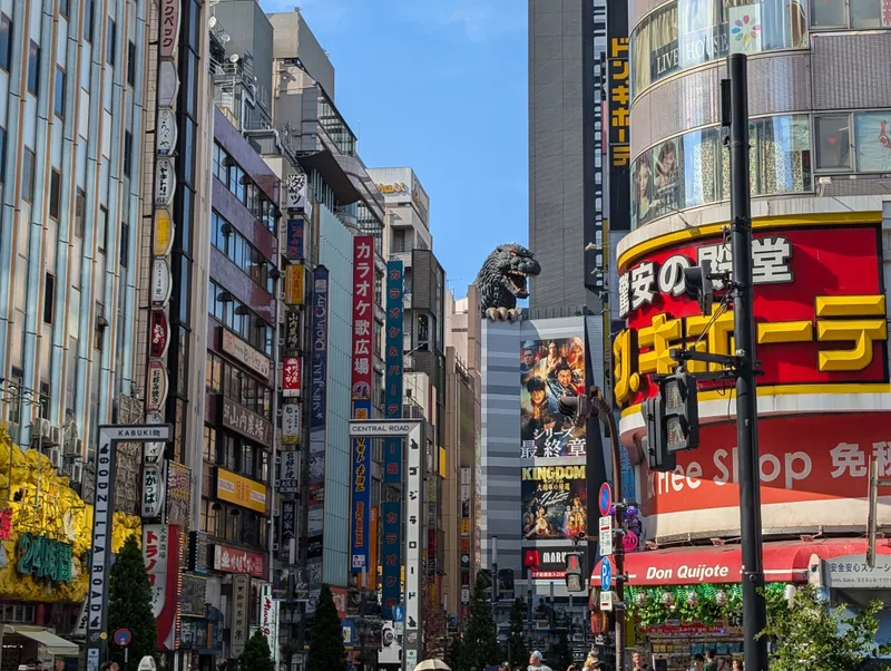 Bustling Shinjuku street in Tokyo, Japan, lined with tall buildings covered in vibrant neon signs and advertisements, with the iconic Godzilla Head visible peeking above a building in the distance.