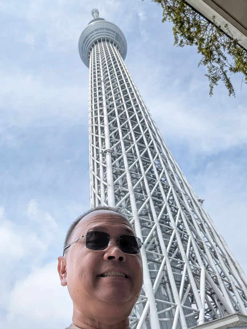 Man in sunglasses smiling at the camera with the Tokyo Skytree towering behind him against a blue sky with light clouds, Tokyo, Japan.