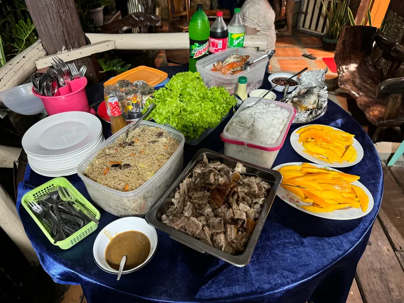 The full spread — rice, noodles, fruit, drinks, and of course the lechon Filipino feast with roasted pig, rice, noodles, mangoes, and drinks on a long table at family gathering in Palawan