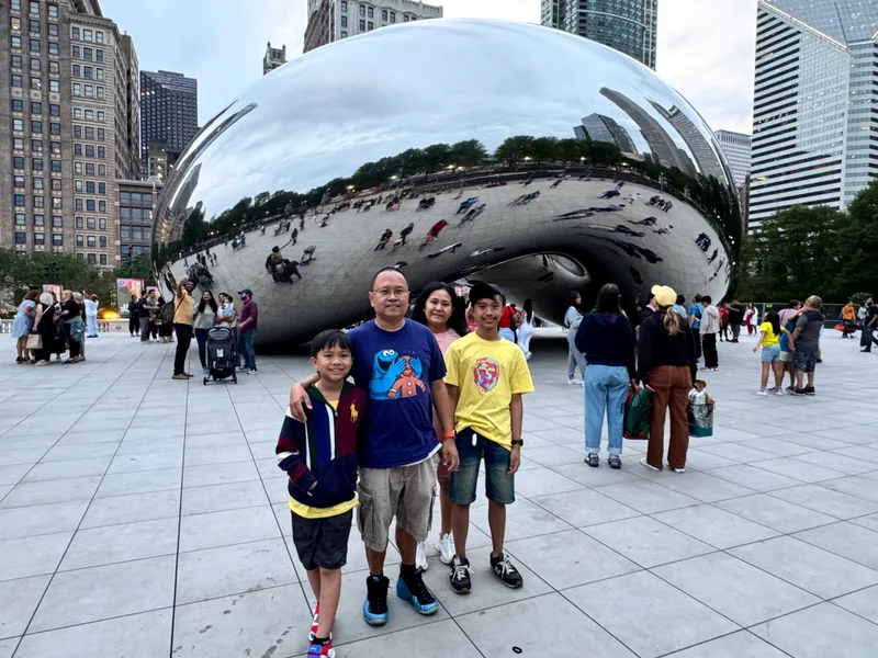 Family of four smiling together in front of Cloud Gate sculpture at Millennium Park, Chicago, Illinois, with city skyline reflected in the sculpture