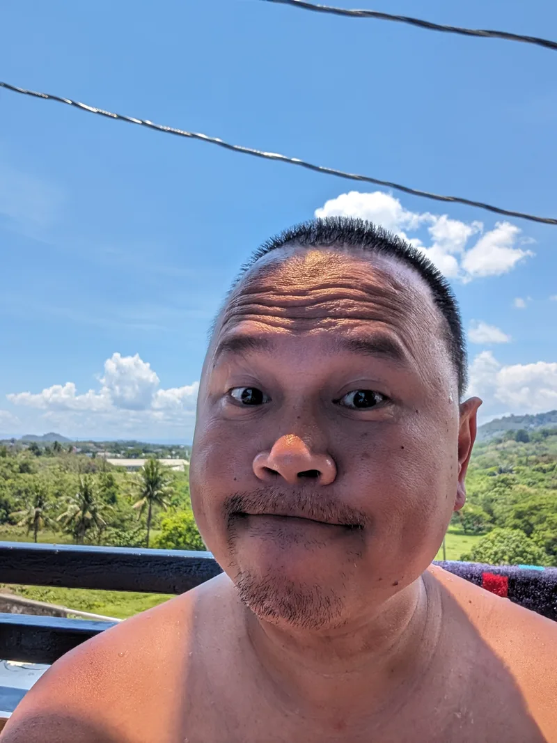 Close-up selfie of a man with a playful expression at Avery Ridge Resort in Pansol, Laguna, with a bright blue sky and green hills.